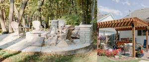 A circular stone patio with 5 adarondak chairs and a built in stone firepit with a retaining wall to protect people from falling off the edge is pictured on the left side of the photo. On the right side of the photo there is a pergola overing a stone laid patio with several chairs, a table, and an outdoor bar.